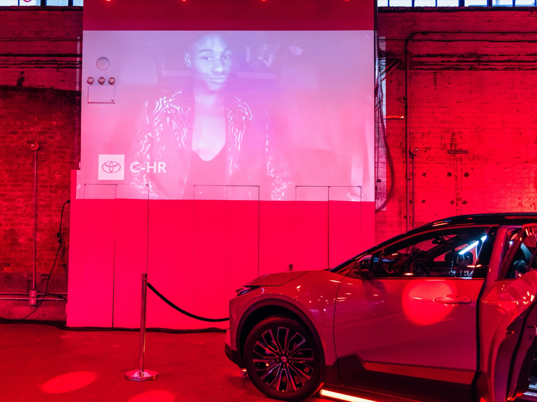 The Toyota C-HR staged on the activation floor at 99 Scott Ave, its door open, with the in-car camera feed projected onto the venue wall behind it under red lighting and a disco ball overhead